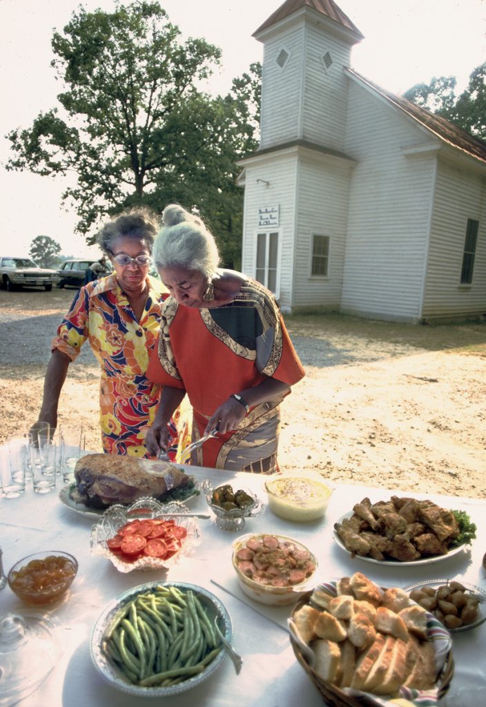 Edna at a table of food with a church visible in the background