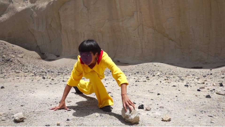 a person with a painted face crouches to grab a rock