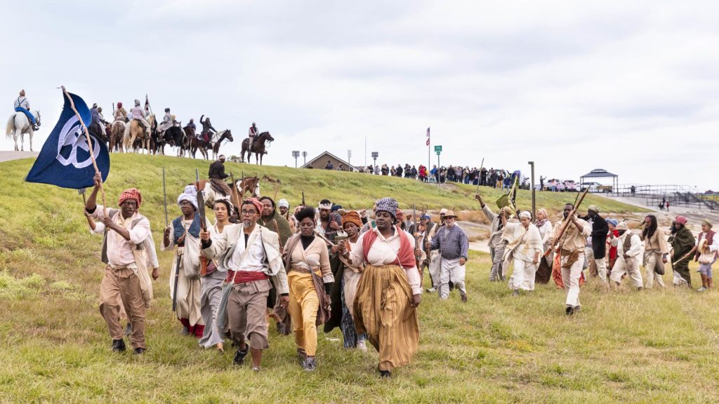 slave revolt reenactors marching while holding a flag and muskets