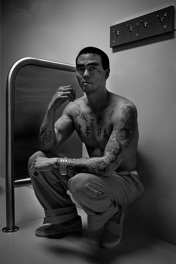 A black and white photo of an Asian male prison squatting with no shirt and visible tattoos and scars.