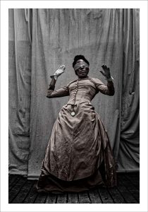 A black woman in victorian dress wearing a blindfold with hands up in the air
