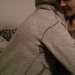 A woman seated on a bed is "embraced" by a woman in a gray hooded sweatshirt.